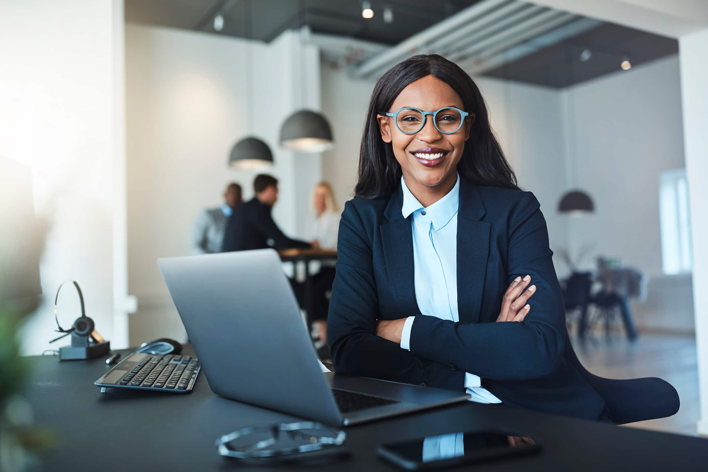 Section image - professional woman smiling while sitting at a desk with a group in the background